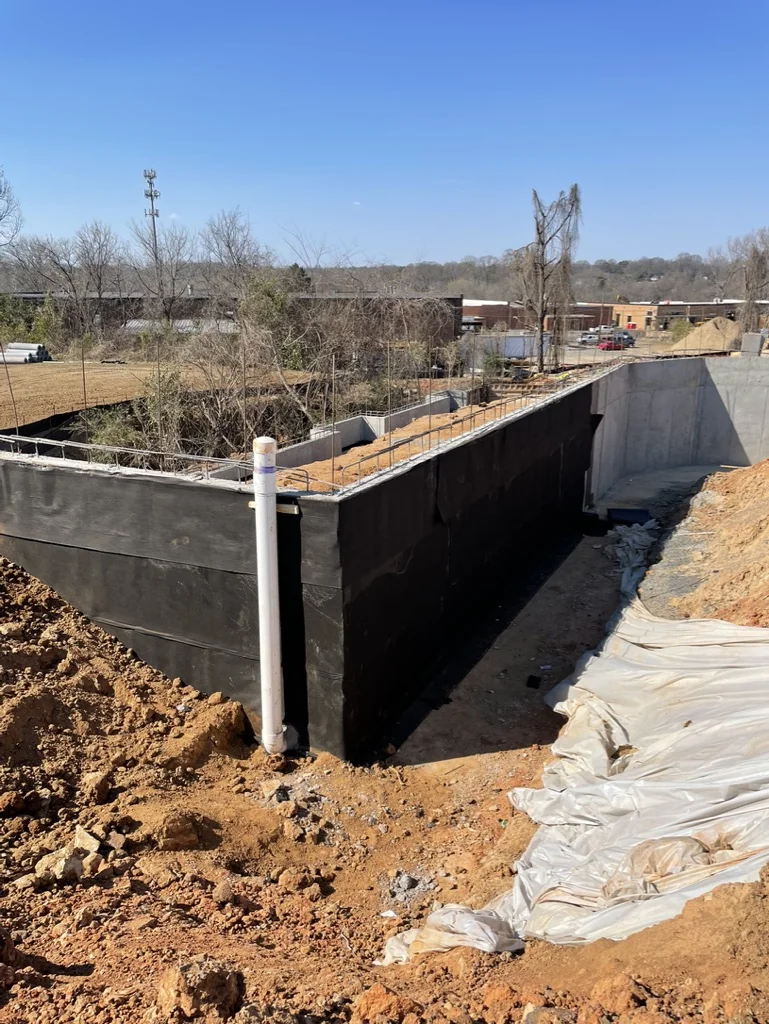 Black material on concrete wall faces beside an open excavation, with a white vertical pipe at the near corner